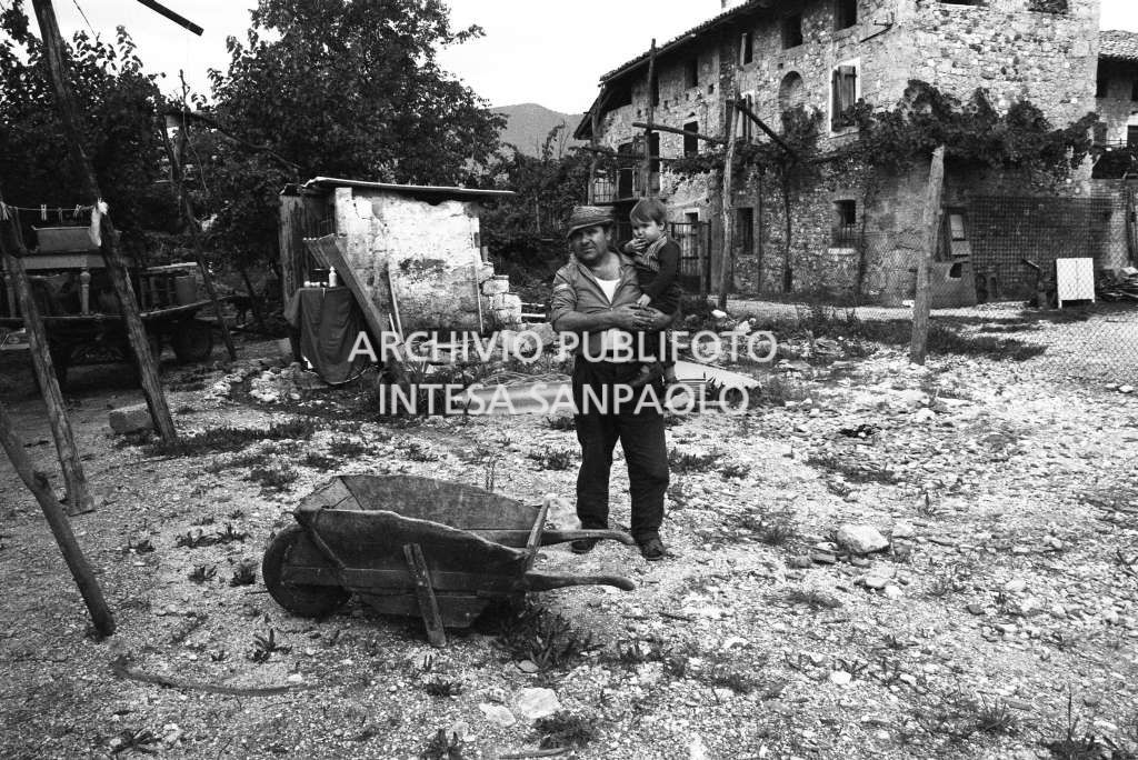 Un uomo ritratto con un bambino in un cortile, visibile alle loro spalle una cascina