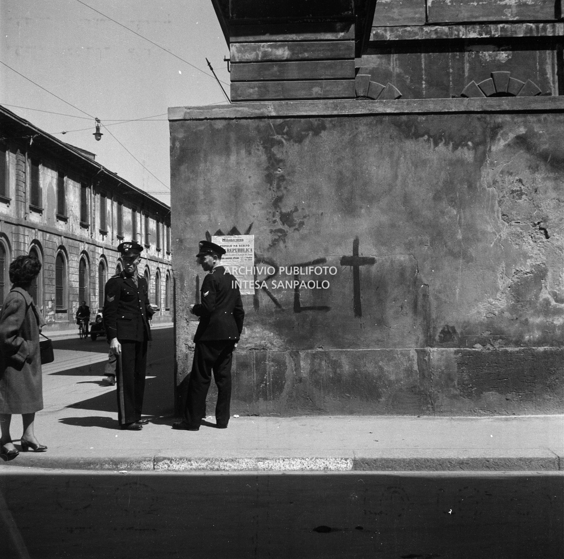 Due carabinieri per strada con una passante commentano presumibilmente la prima pagina della seconda edizione del quotidiano Milano-Sera, affissa a un muro, che titola: "Il popolo ha scelto. È già Repubblica"