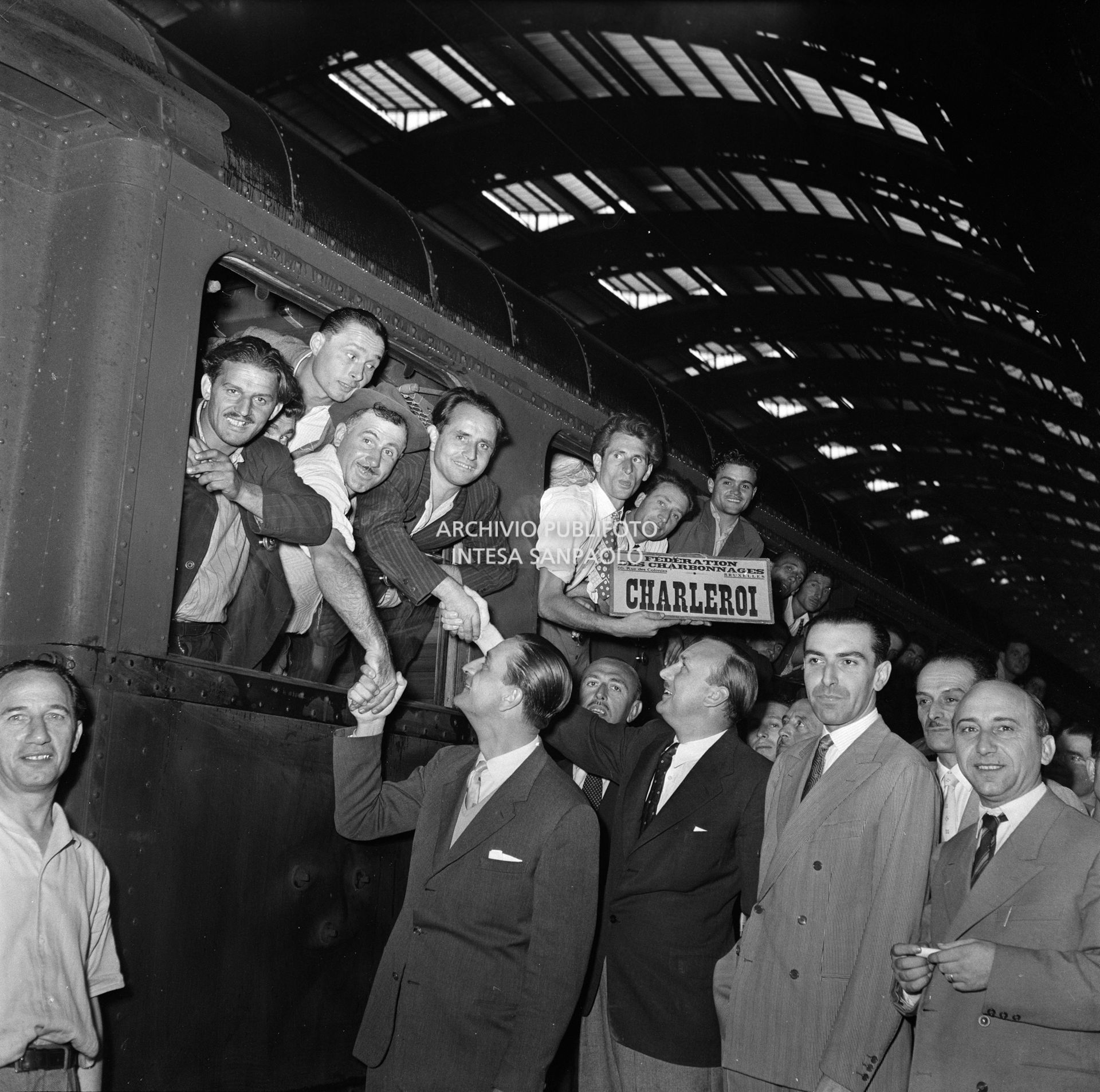 Alfred Robens, ministro inglese del Lavoro, tra gli altri, saluta gli emigranti, affacciati al vagone ferroviario di un treno, in partenza per le miniere del Belgio, dalla Stazione Centrale di Milano. Uno di loro tiene in mano il cartello: "Federation des charbonnages 66, rue des Colonies, Bruxelles. Campine"