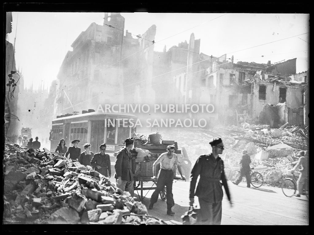 Scorcio di corso Vittorio Emanuele II e di piazza San Carlo a Milano, all'indomani dei bombardamenti: militari, civili, un religioso, e un uomo che tira un carretto, camminano in fila indiana a lato della strada dove sono state addossate le macerie degli edifici distrutti durante gli attacchi aerei. Un tram tipo 1928, linea 3 per Lambrate, avanza nella via. Sullo sfondo il Duomo