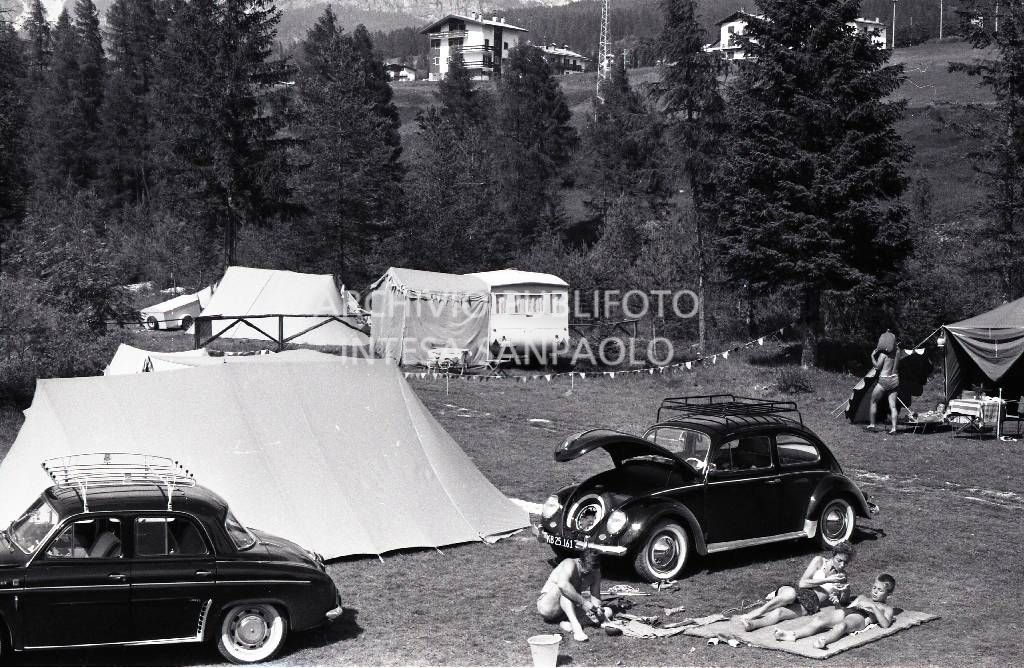 Turisti in campeggio a Cortina d'Ampezzo sdraiati in un prato a fianco a un Maggiolino Volkswagen ; sulla sinistra una Renault Dauphine/Ondine<br>706765/64