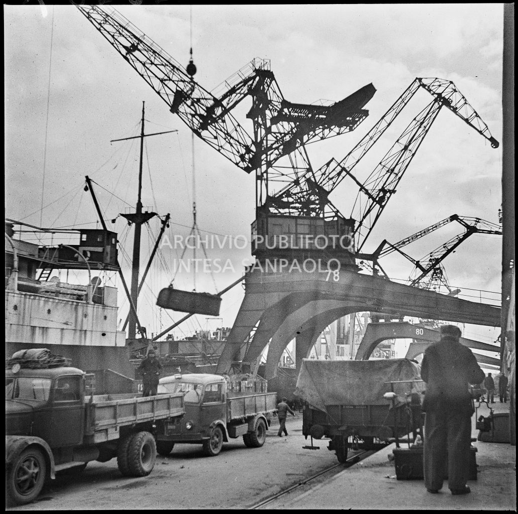 Carico merce per l'esportazione al porto di Genova. A sinistra due autocarri in sosta sulla banchina e sullo sfondo numerose gru portuali<br>23267