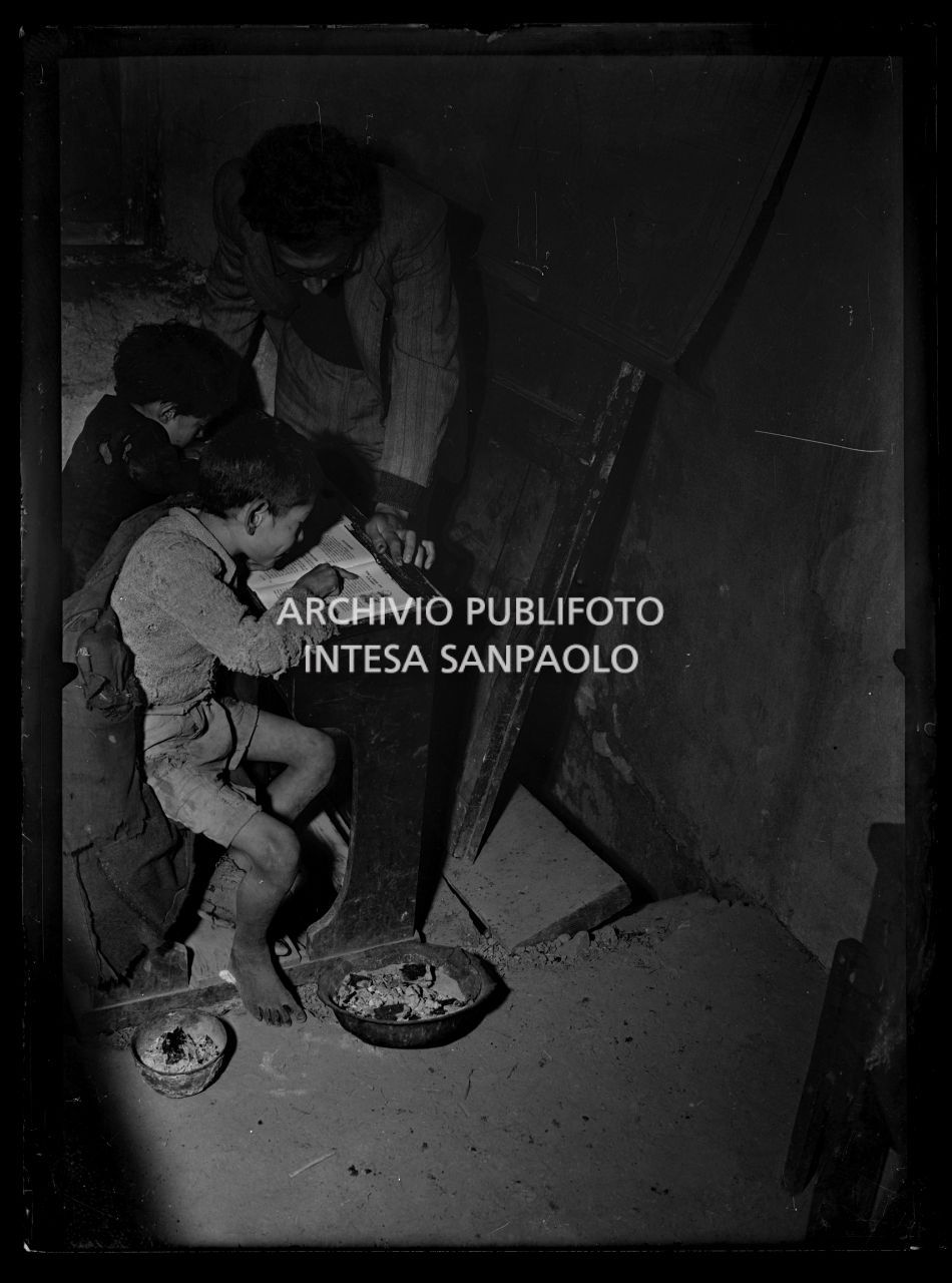 Africo, due bambini scalzi seduti a un banco di scuola. Il maestro li segue nella lettura
