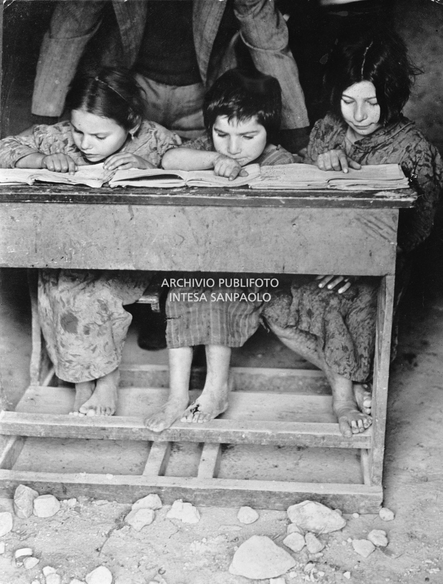 Africo, bambine scalze sedute a un banco di scuola intente alla lettura; alle loro spalle il maestro segue l'esercitazione