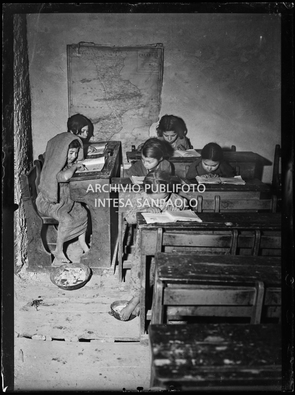 Africo, bambini scalzi seduti ai banchi di scuola; alla parete è affissa la carta geografica della Calabria