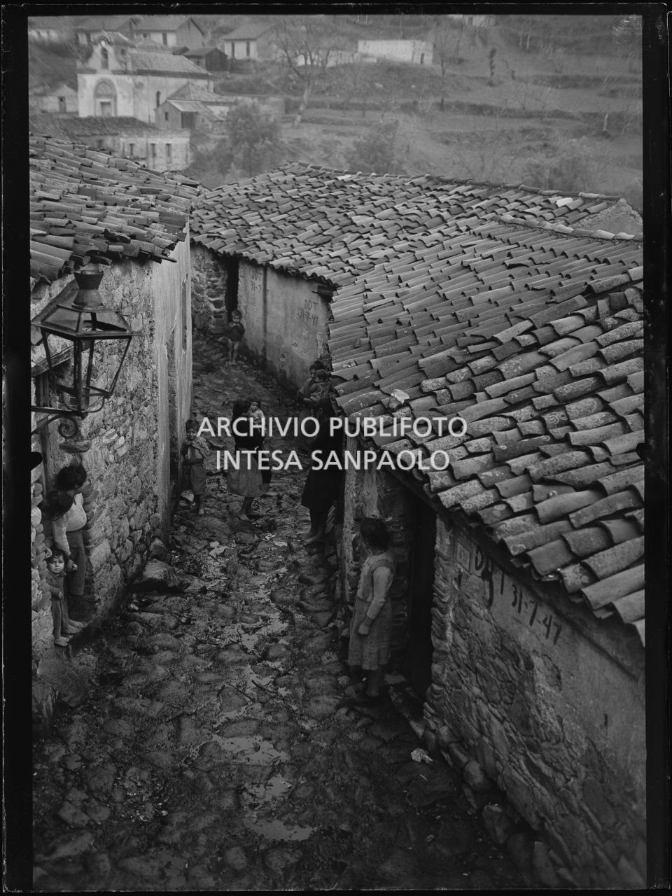 Africo, scorcio di un viottolo tra le case del paese con bambini e donne; sullo sfondo la chiesa