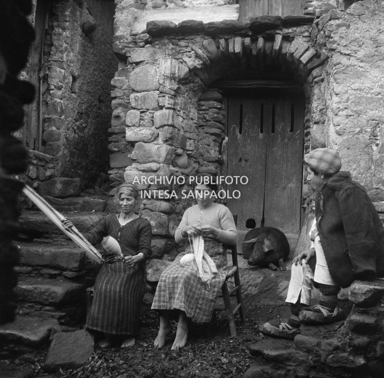 Africo, due donne sedute, una fila la lana e l'altra lavora a maglia osservate da un ragazzo con indosso le cioce, calzature tradizionali; alle loro spalle un maiale