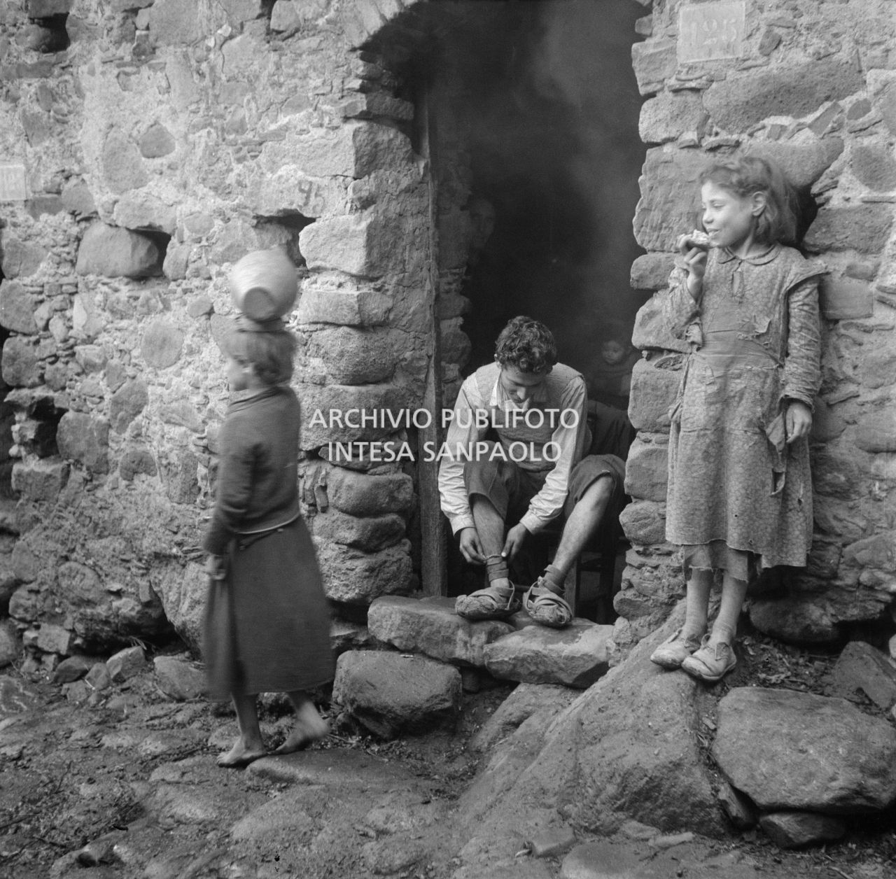 Africo, un ragazzo calza delle cioce, calzature tradizionali, seduto sulla soglia di casa; una ragazzina  mangia del pane e un'altra si avvia con una giara sul capo