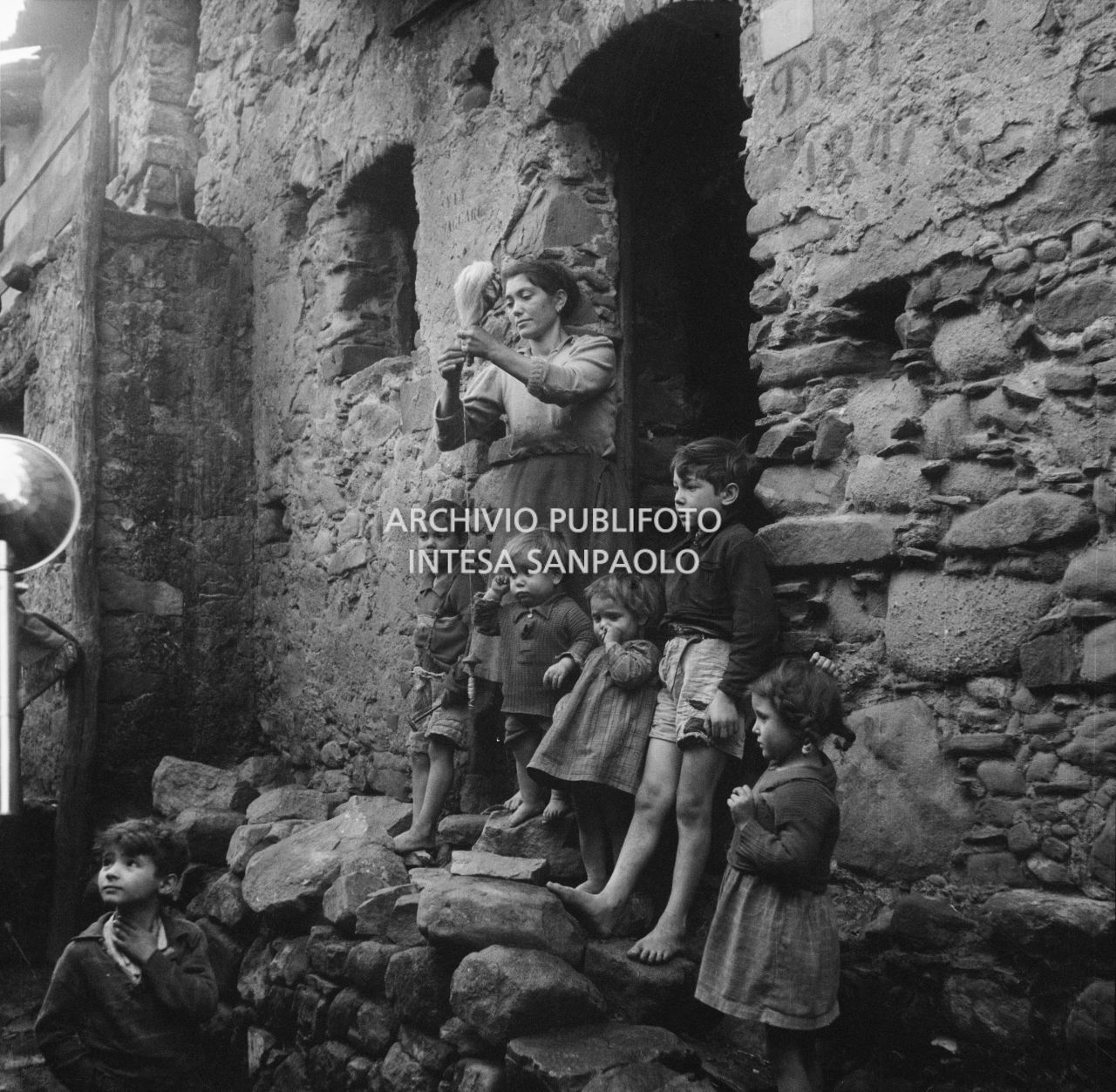 Africo, una donna con in mano un fuso di filato ritratta sulla soglia di casa insieme a otto bambini