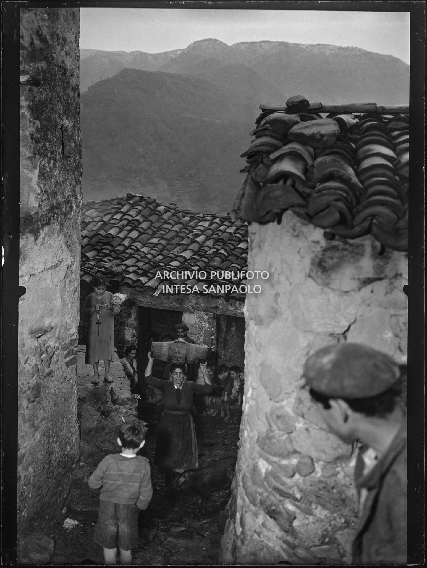 Africo, una donna risale un vicolo del paese trasportando una botte sulla testa, tra bambini, un maiale e altri abitanti