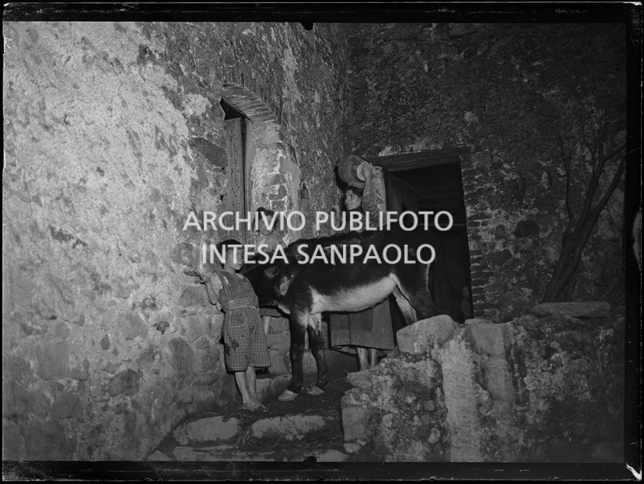 Africo, una donna, con in testa una giara, due bambini e un asino all'esterno di una casa