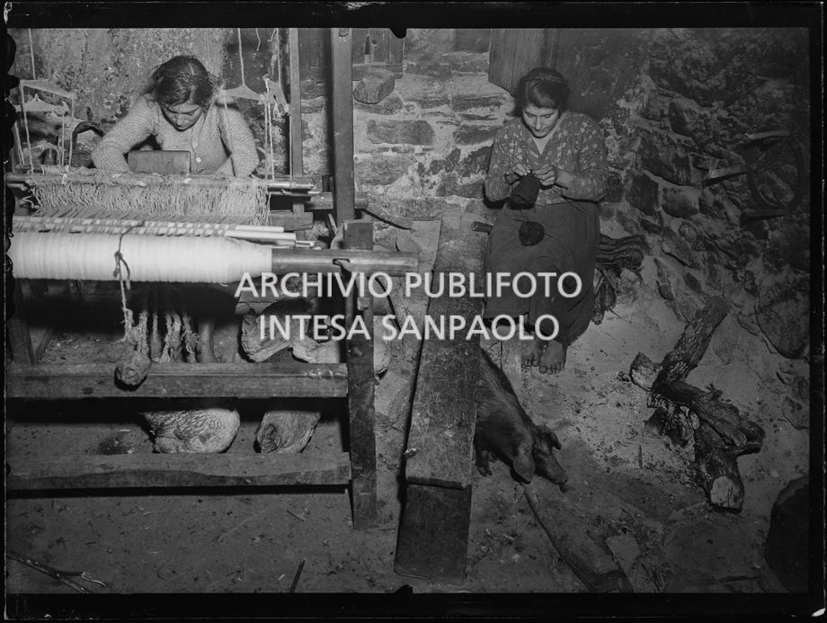 Africo, interno con due donne: una al telaio e l'altra lavora a maglia tra galline che razzolano e un maiale