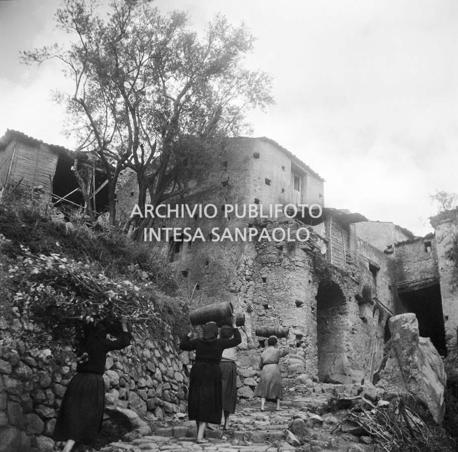 Africo, donne trasportano sulla testa botti di legno e fascine di legna salendo una scalinata del paese