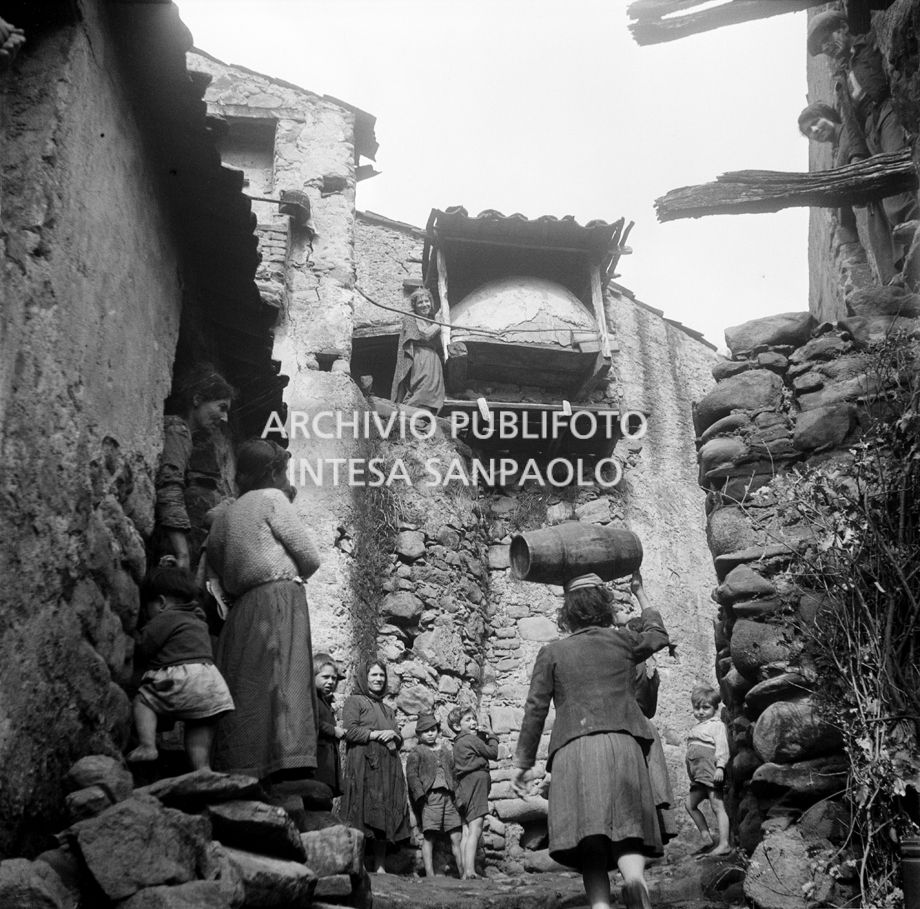 Africo, scorcio di un vicolo con bambini e donne: una trasporta una botte di legno sulla testa