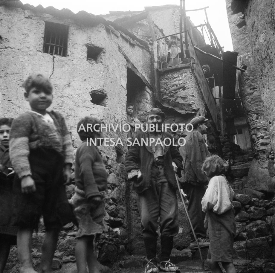 Africo, scene di vita quotidiana in un vicolo: al centro una donna trasporta una botte di legno sulla testa