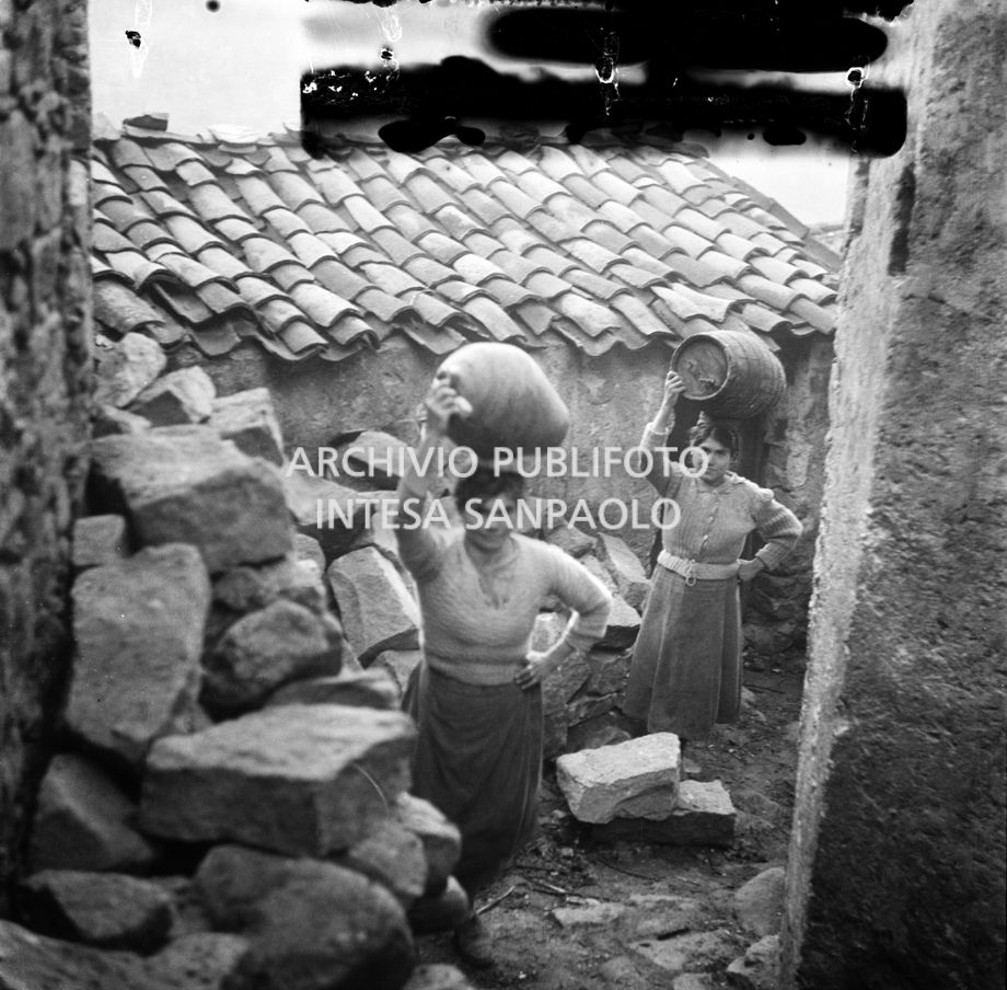 Africo, due ragazze con botti di legno e giare sulla testa ritratte in un angolo di paese