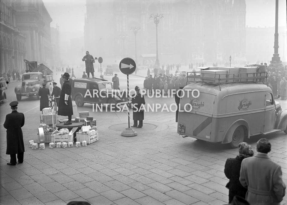 Befana del vigile in piazza Duomo a Milano: alla base della pedana,  dalla quale viene regolato il traffico, sono depositati i doni per i vigili della città, sullo sfondo il furgoncino della Publifoto sul cui tetto è appostato un fotografo per riprendere l'evento<br>63284