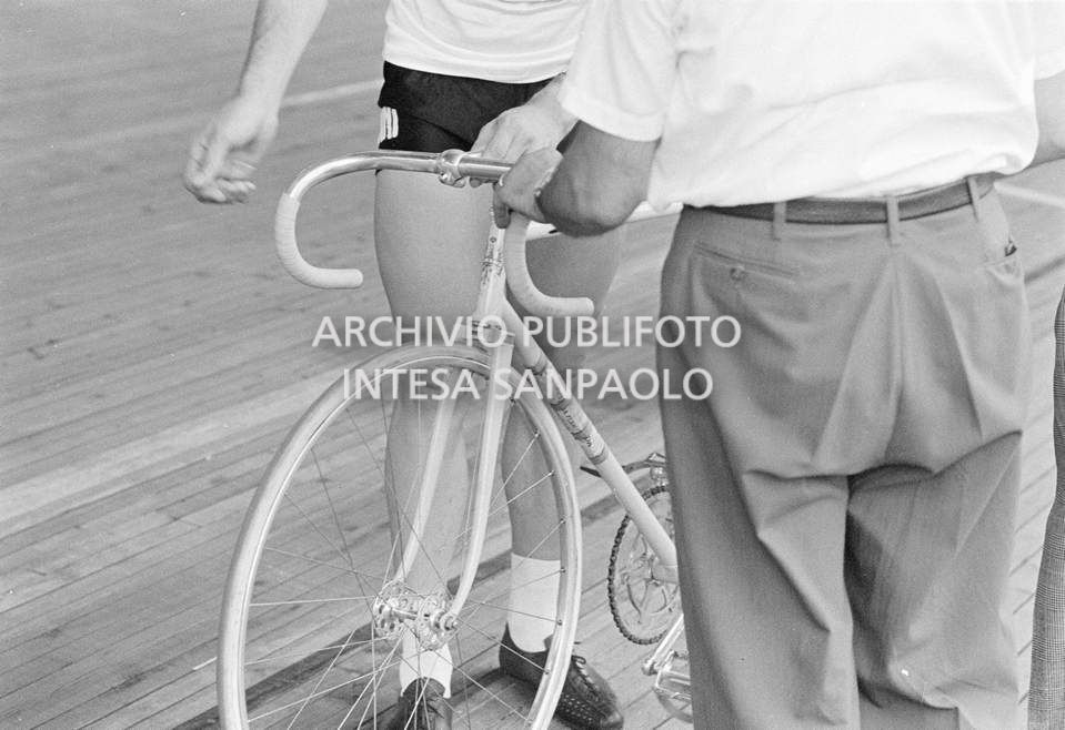 Roger Rivière e il meccanico Giuseppe De Grandi, detto Pinella, dei quali vediamo solo le gambe, con la bicicletta preparata da quest'ultimo, in pista per gli allenamenti in vista della competizione per il record dell'ora