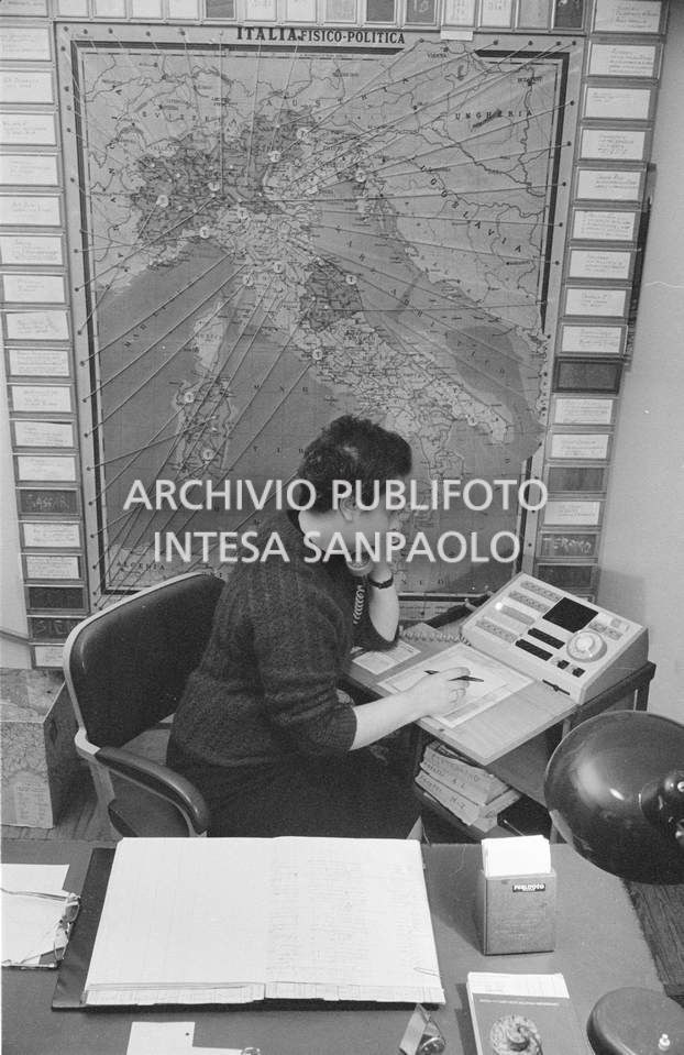 Addetta al centralino dell'agenzia Publifoto nella sede di via Bramante a Milano; dietro di lei una carta fisico politica dell'Italia con dei segnaposto numerati