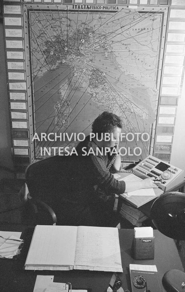 Addetta al centralino dell'agenzia Publifoto nella sede di via Bramante a Milano; dietro di lei una carta fisico politica dell'Italia con dei segnaposto numerati