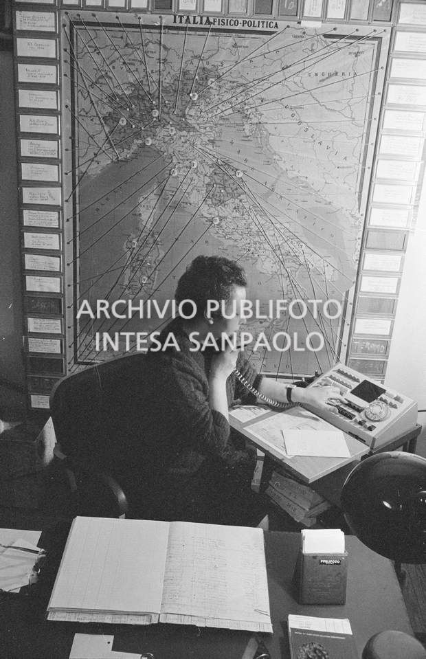 Addetta al centralino dell'agenzia Publifoto nella sede di via Bramante a Milano; dietro di lei una carta fisico politica dell'Italia con dei segnaposto numerati