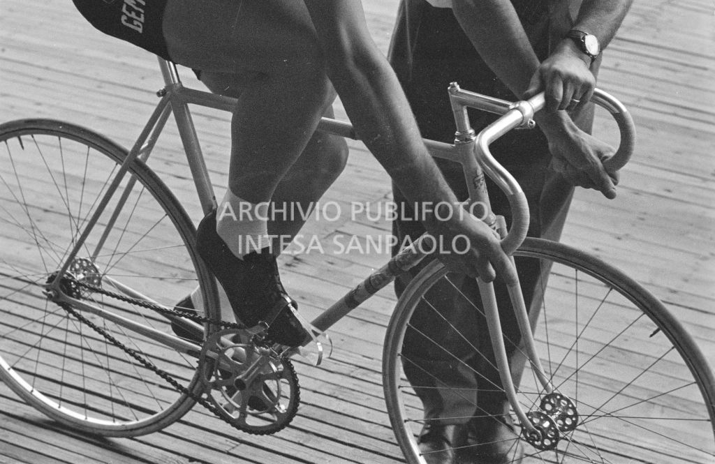 Roger Rivière e il meccanico Giuseppe De Grandi, detto Pinella, dei quali vediamo solo le gambe, con la bicicletta preparata da quest'ultimo, in pista per gli allenamenti in vista della competizione per il record dell'ora