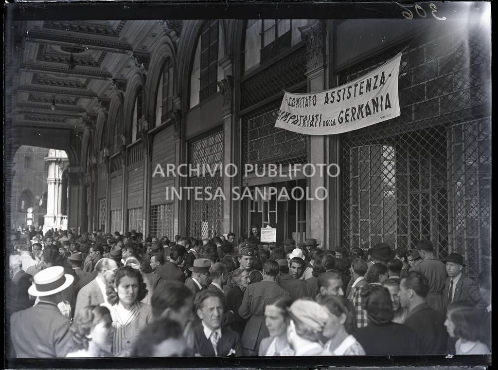 Folla di gente sotto i portici settentrionali di piazza del Duomo, visibile su una delle vetrine lo striscione che reca la scritta: Comitato Assistenza Rimpatriati dalla Germania<br>10306