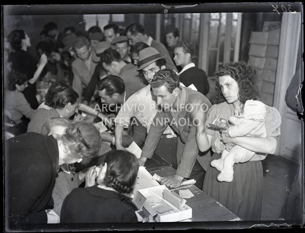 Una donna con un bambino in braccio si rivolge al banco del Comitato Assistenza Rimpatriati dalla Germania; visibili in secondo piano numerosi uomini<br>10307