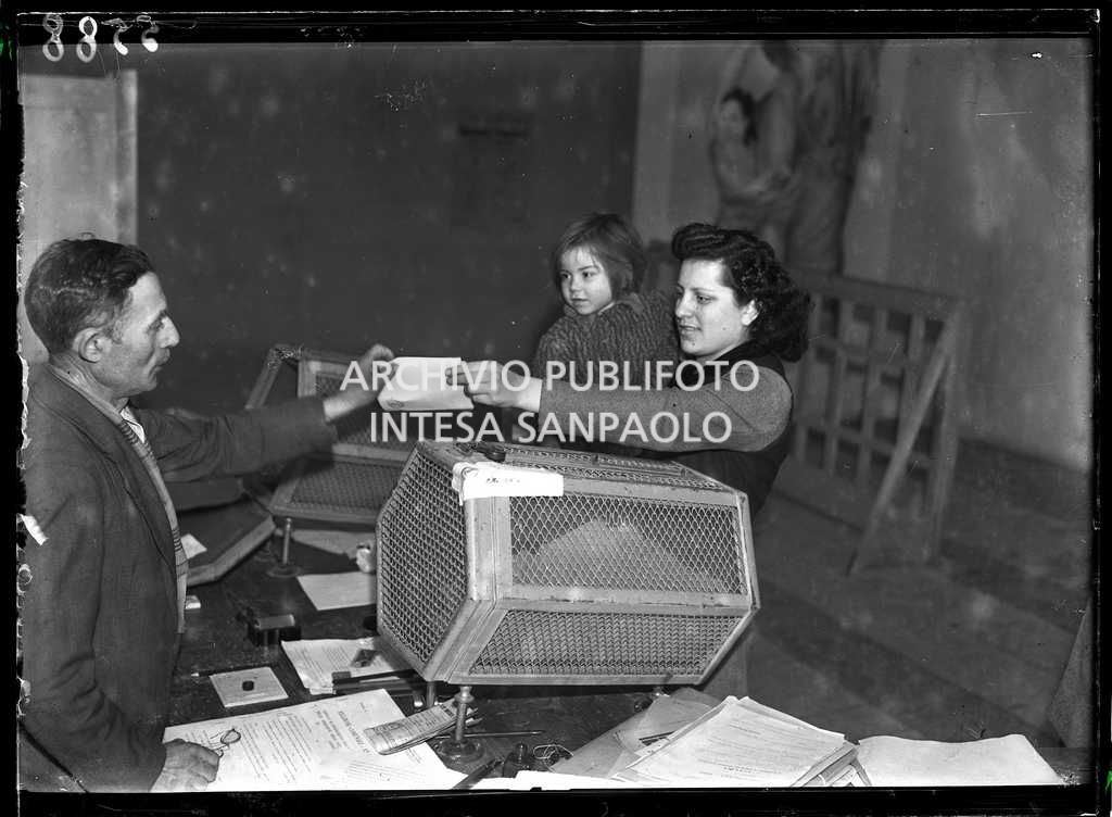 Una donna, con un bambino in braccio, consegna il certificato elettorale al seggio in occasione delle elezioni comunali nel Lodigiano, le prime elezioni con suffragio universale in Italia