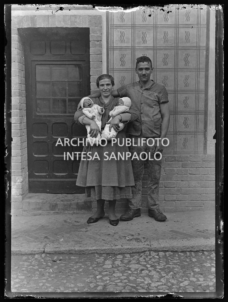 Una coppia, con due neonati in fasce, ritratta davanti alla porta di casa