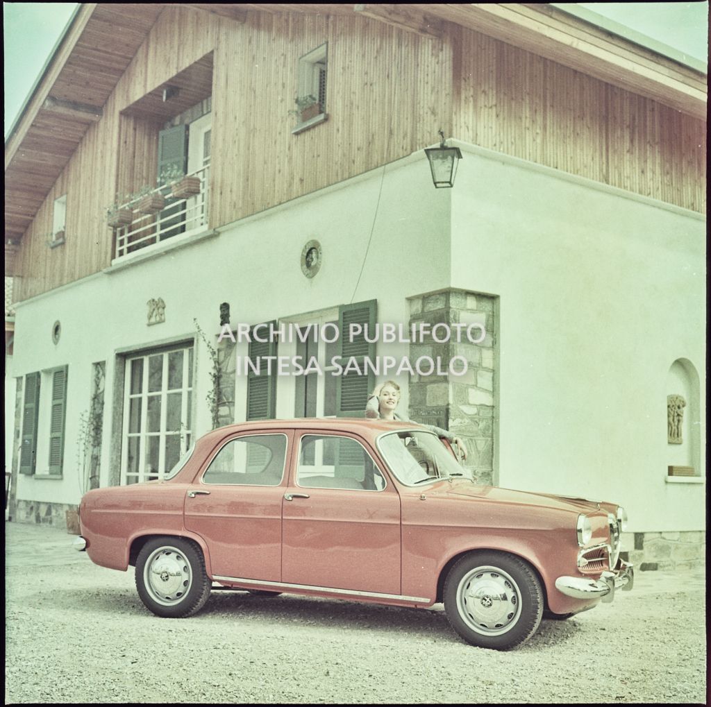 Lato destro e muso di una Alfa Romeo Giulietta berlina di colore rosso davanti ad un'abitazione con una modella in piedi dal lato guidatore