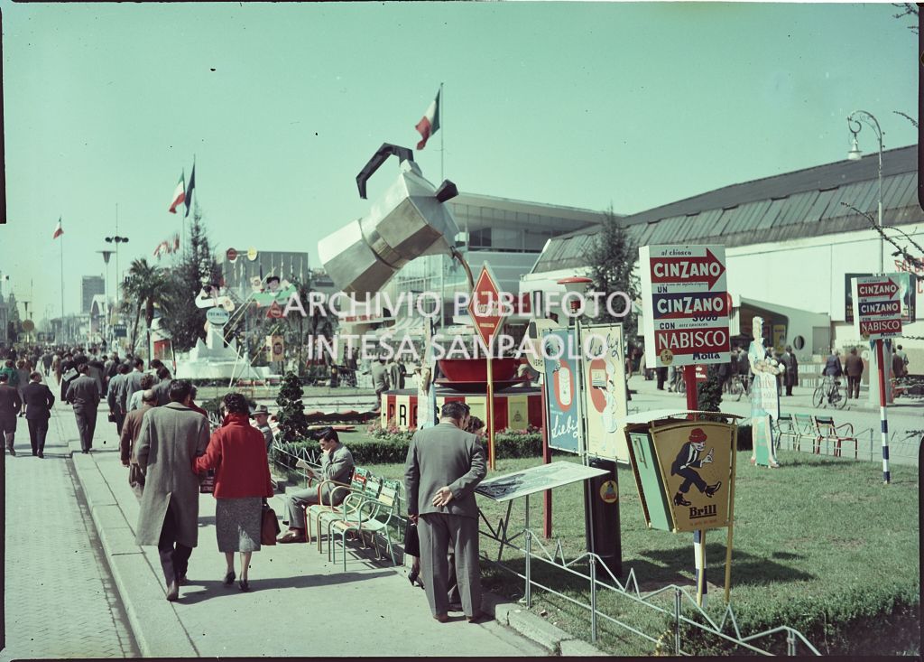 Vista esterna della Fiera Campionaria di Milano del 1955 con visitatori. In secondo piano un'enorme caffettiera versa il caffè un una grande tazzina