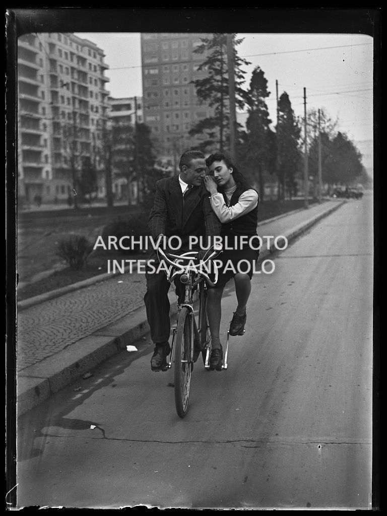 Un uomo e una donna pedalano su un particolare modello di bicicletta in piazza della Repubblica a Milano
