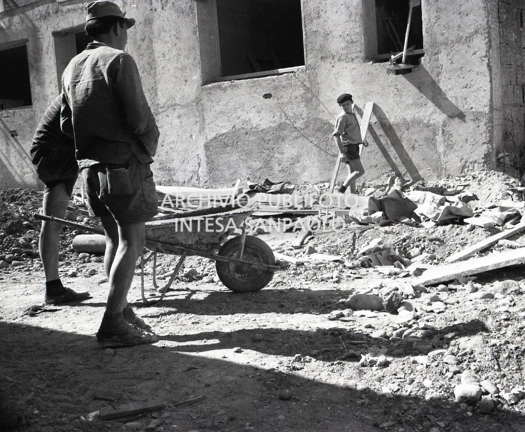 Ragazzino al lavoro in un cantiere edile: in primo piano due altri manovali con carriola<br>721238/4