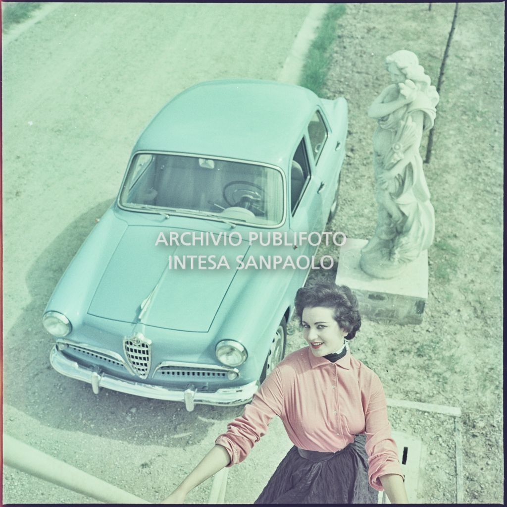 Alfa Romeo Giulietta berlina azzurra esposta in un giardino con una modella in primo piano su una scala