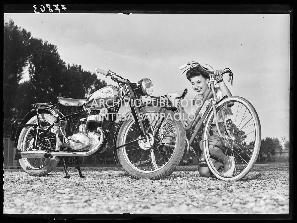 Una donna posa accanto a una moto, reggendo una bicicletta