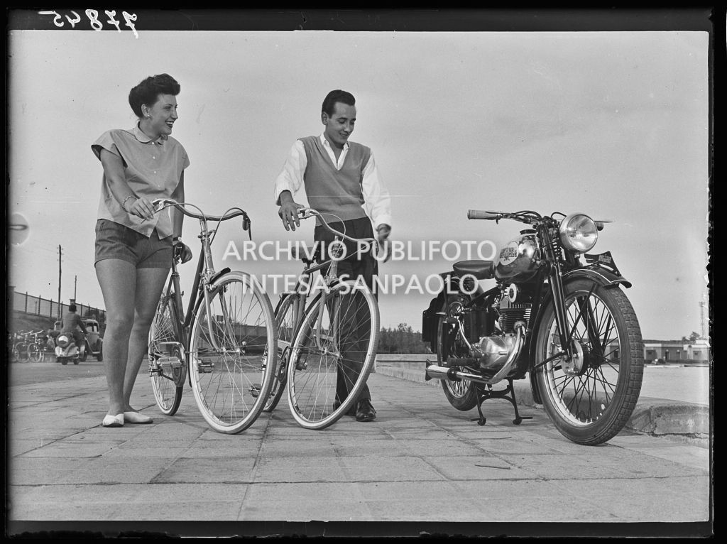 Un uomo e una donna, reggendo le loro biciclette, ammirano una motocicletta parcheggiata