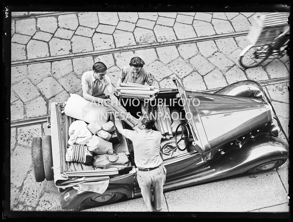 Tre persone, riprese dall'alto, caricano generi alimentari impacchettati su un'automobile decappottabile