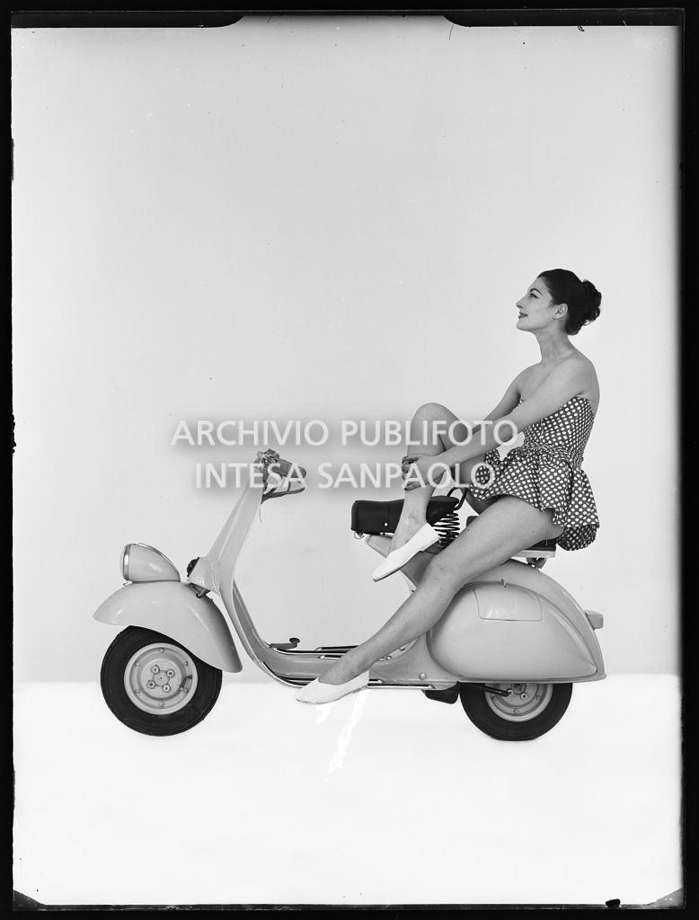 Una modella posa, su limbo bianco, seduta sul sellino passeggero di una Vespa 125 VN2T