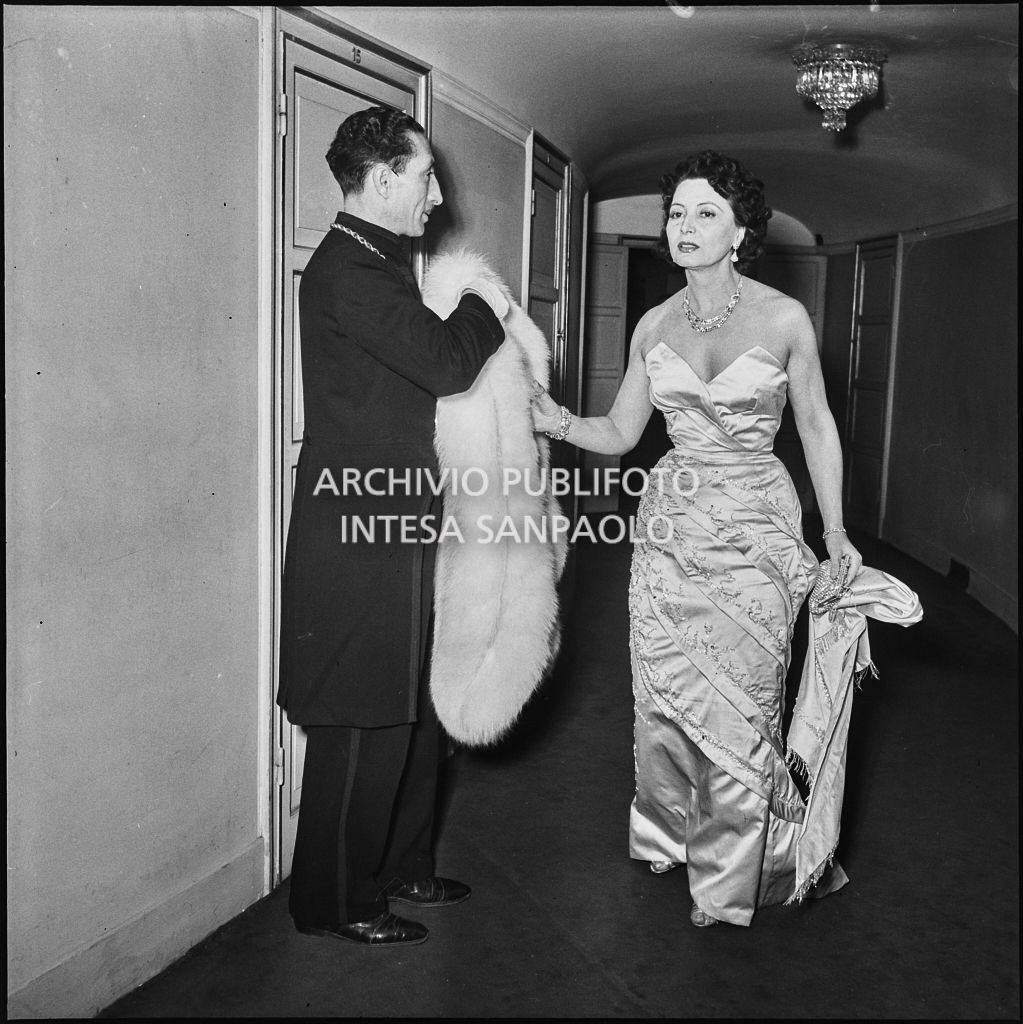 Una maschera assiste la baronessa Bianca Maria Hubner al Teatro alla Scala in occasione della serata inaugurale della stagione lirica 1953-1954 con l'opera "La Wally", di Alfredo Catalani, diretta da Carlo Maria Giulini, con la regia di Tatiana Pavlova