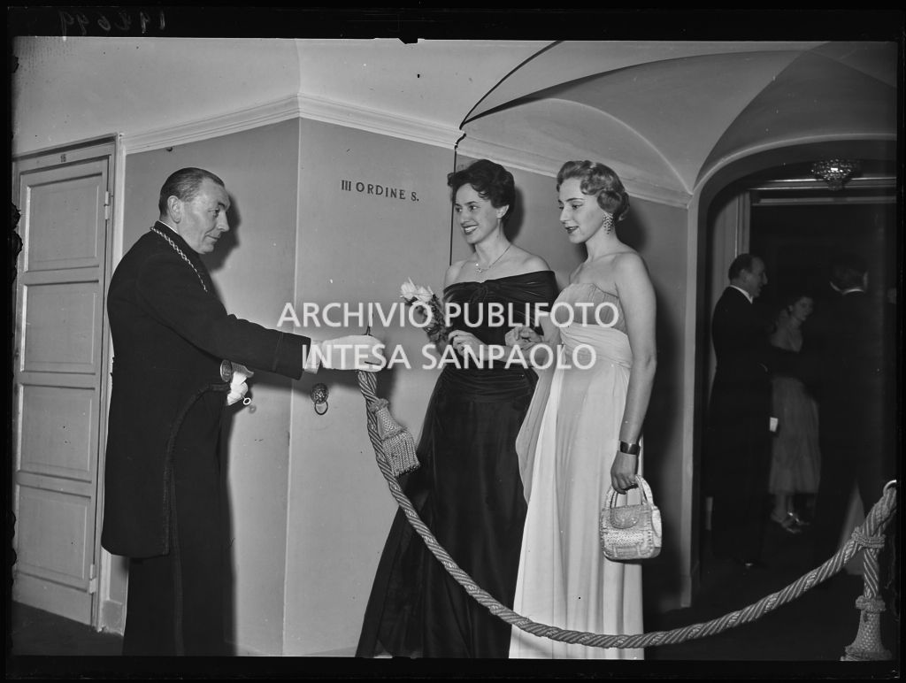 Una maschera del Teatro alla Scala accoglie nel corridoio di accesso ai palchi, Marcella De Girolami, moglie di Carlo Maria Giulini e Emanuela di Castelbarco Toscanini (a destra) la sera dell'inaugurazione della stagione lirica 1953-1954 con l'opera "La Wally", di Alfredo Catalani, diretta da Carlo Maria Giulini, con la regia di Tatiana Pavlova