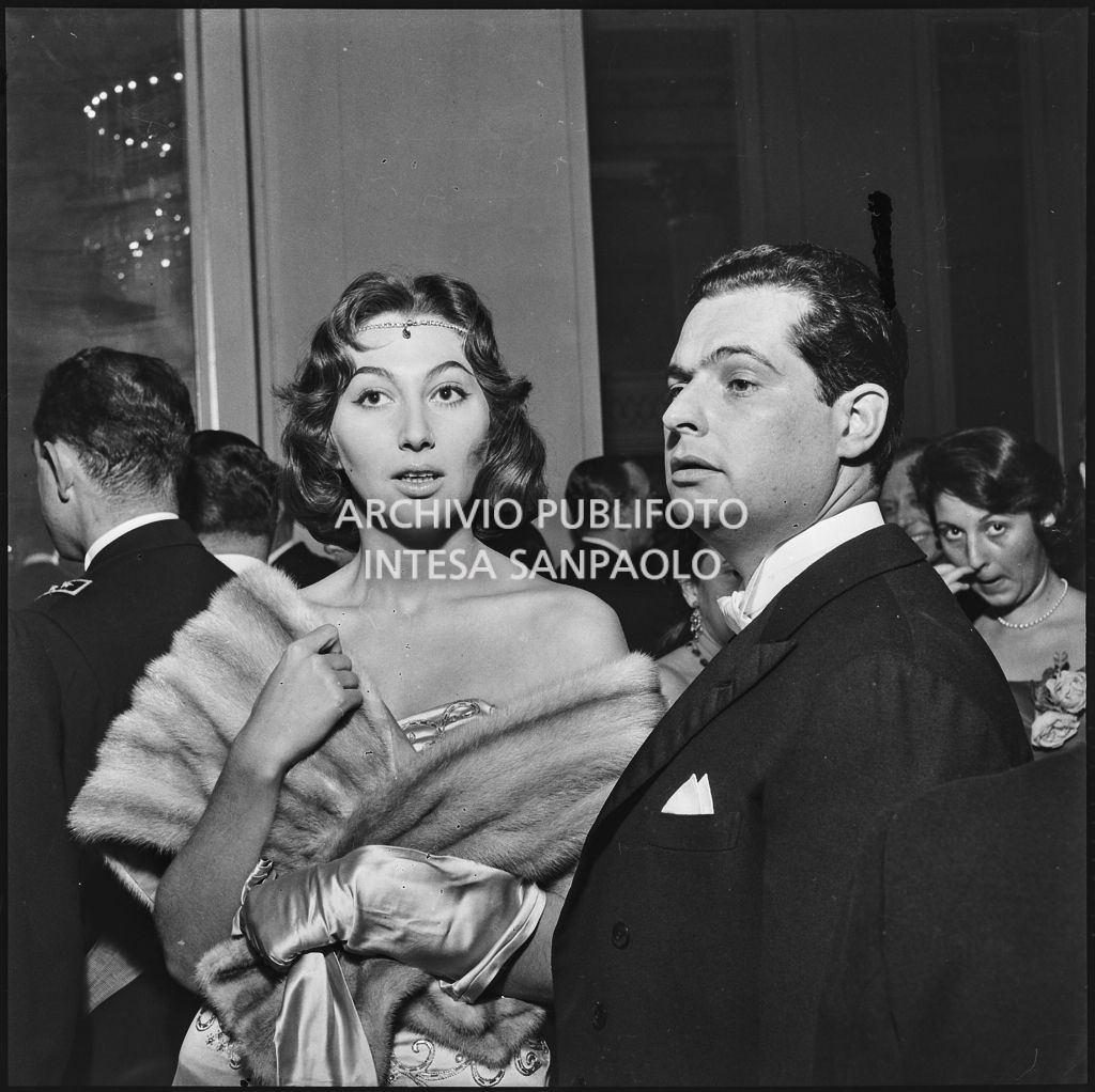 Pini Carnevale in compagnia di un uomo nel foyer del Teatro alla Scala in occasione della serata inaugurale della stagione lirica 1957-1958 con l'opera "Un ballo in maschera", di Giuseppe Verdi, diretta da Gianandrea Gavazzeni e la regia di Margherita Wallmann