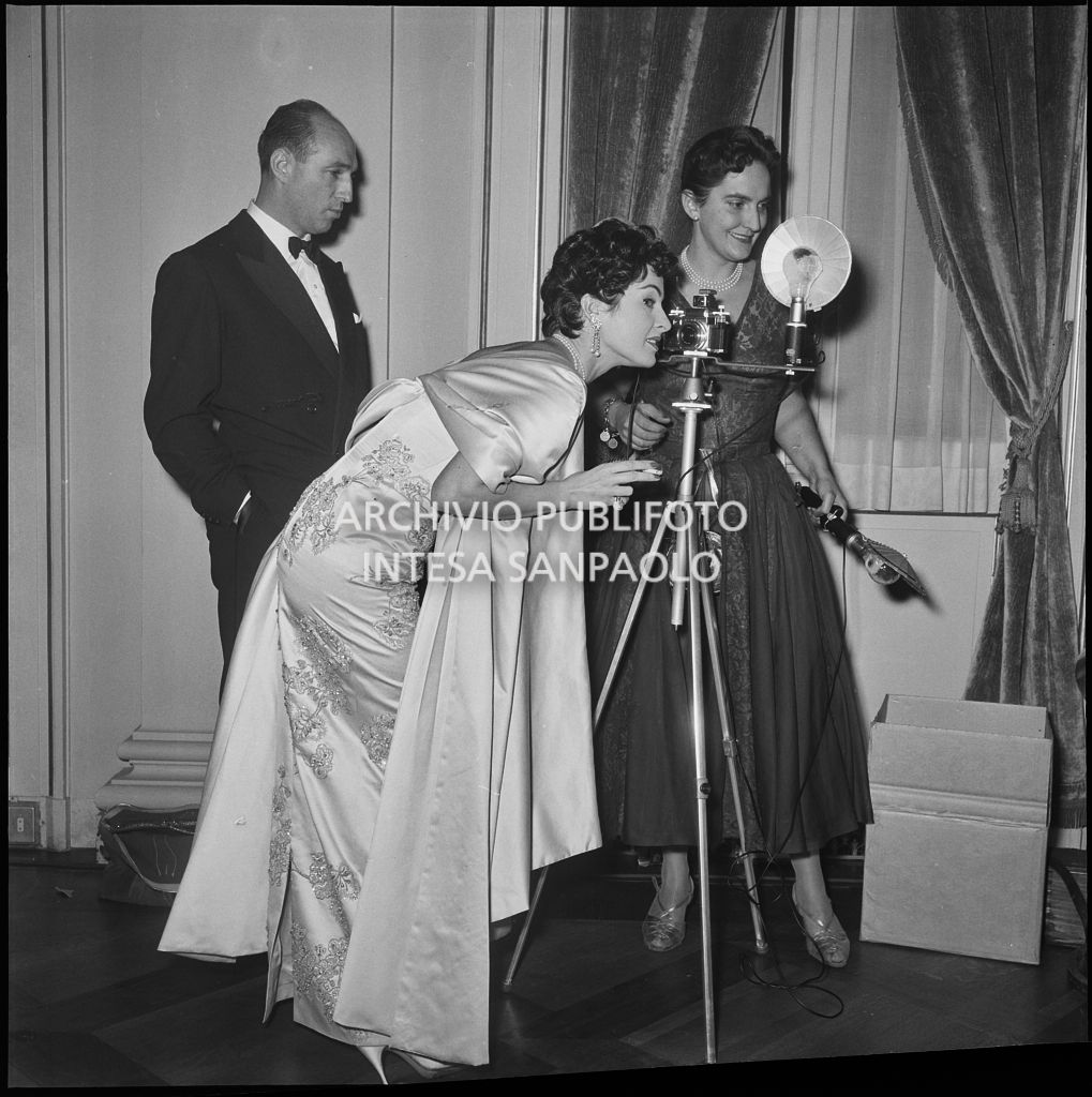 Silvana Pampanini, piegata su una macchina fotografica, e Ludovica Doria, al Teatro alla Scala, in occasione della serata inaugurale della stagione lirica 1956-1957 con l'opera "Aida" di Giuseppe Verdi, diretta da Antonino Votto, con la regia di Franco Enriquez