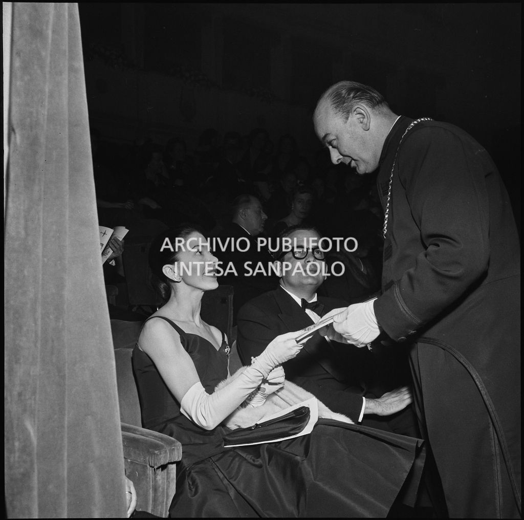 Margot Fonteyn, seduta in platea con il marito Roberto Arias, riceve il libretto d'opera da una maschera alla Piccola Scala in occasione della serata inaugurale della stagione lirica 1956-1957 con l'opera "La cecchina ossia la buona figliola" di Niccolò Vito Piccinni, diretta da Nino Sanzogno, con la regia di Franco Zeffirelli