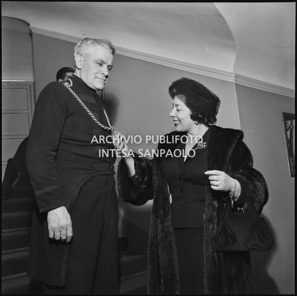 Il mezzosoprano Giulietta Simionato ammira il medaglione indossato da una maschera del Teatro alla Scala in occasione della serata inaugurale della stagione lirica 1957-1958 con l'opera di Giuseppe Verdi "Un ballo in maschera", diretta da Gianandrea Gavazzeni e con la regia di Margherita Wallmann