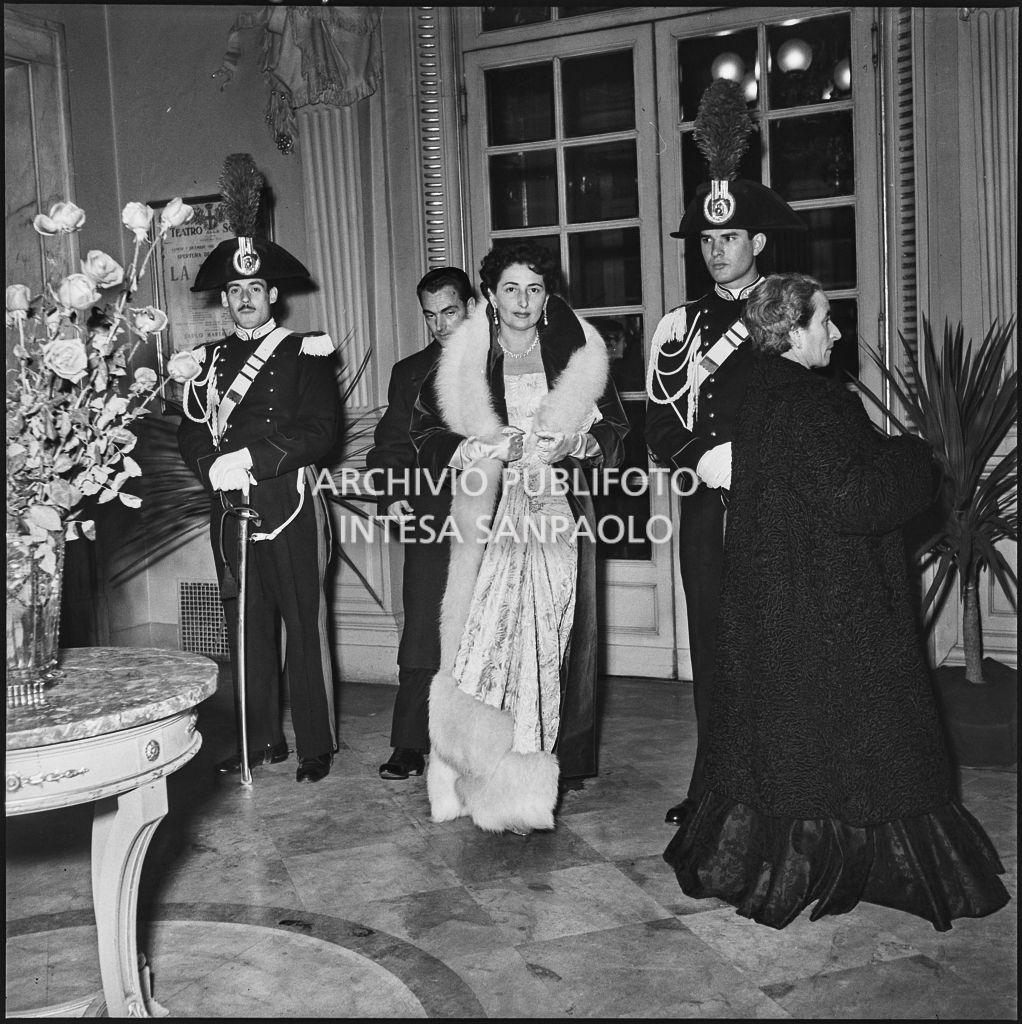 I coniugi Campatelli al loro ingresso nel foyer del Teatro alla Scala, tra due carabinieri, in occasione della serata inaugurale della stagione lirica 1953-1954 con l'opera "La Wally", di Alfredo Catalani, diretta da Carlo Maria Giulini, con la regia di Tatiana Pavlova