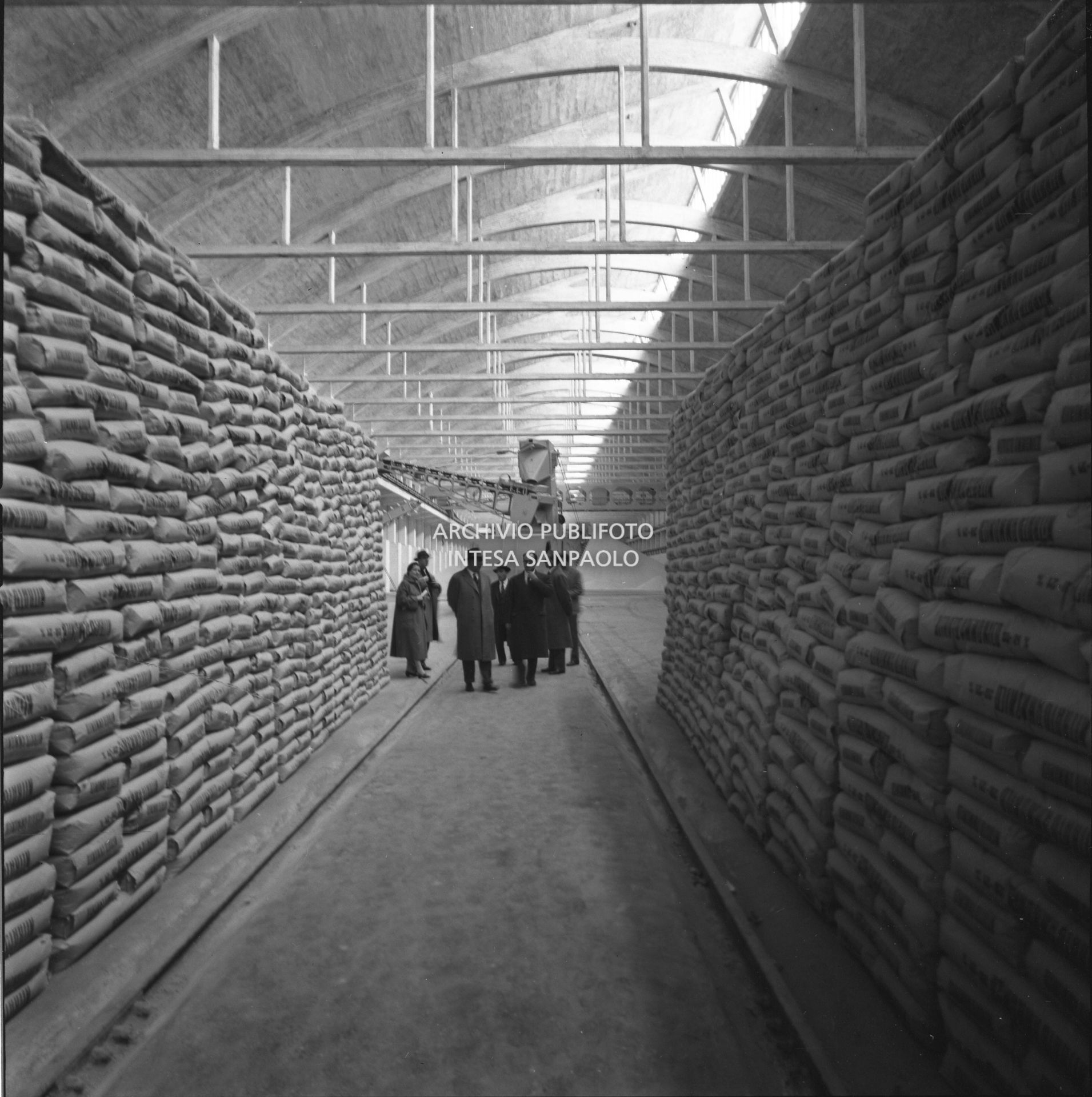 Interno del magazzino: ai lati sacchi impilati, al centro in fondo visitatori con alle spalle macchinario insacchettatore<br>441576