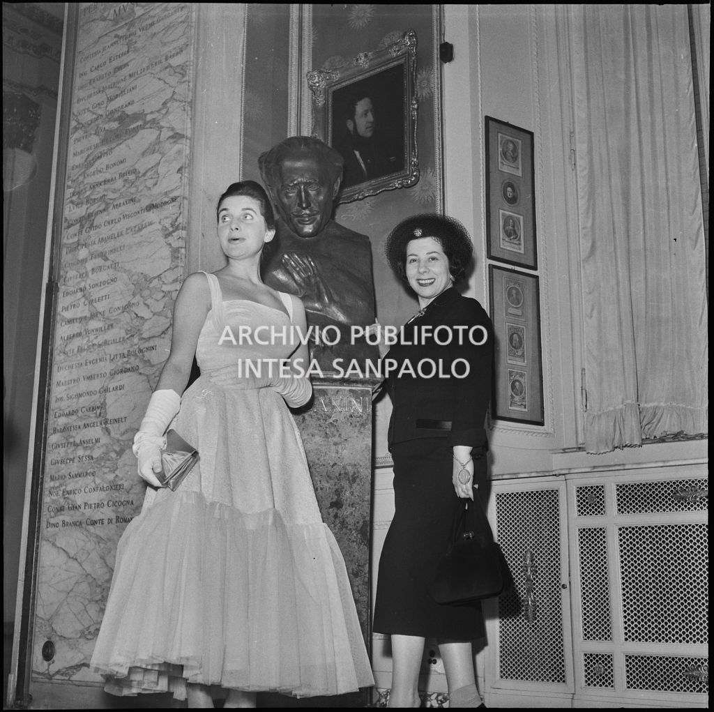 Il mezzosoprano Giulietta Simionato e Antonia Magri, nipote di Antonio Ghiringhelli, in posa davanti al busto di Toscanini al Teatro alla Scala in occasione della serata inaugurale della stagione lirica 1957-1958, aperta con l'opera "Un ballo in maschera", di Giuseppe Verdi, diretta da Gianandrea Gavazzeni con la regia di Margherita Wallmann