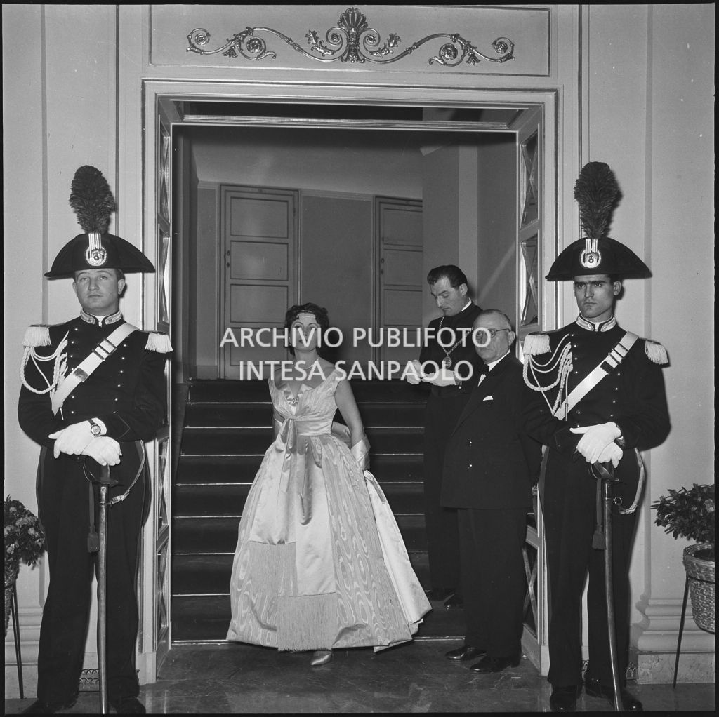 La baronessa Bianca Maria Hubner in posa tra maschere e carabinieri nel foyer del Teatro alla Scala in occasione della serata inaugurale della stagione lirica 1958-1959 con l'opera "Turandot", di Giacomo Puccini, diretta da Antonino Votto con la regia di Margherita Wallmann