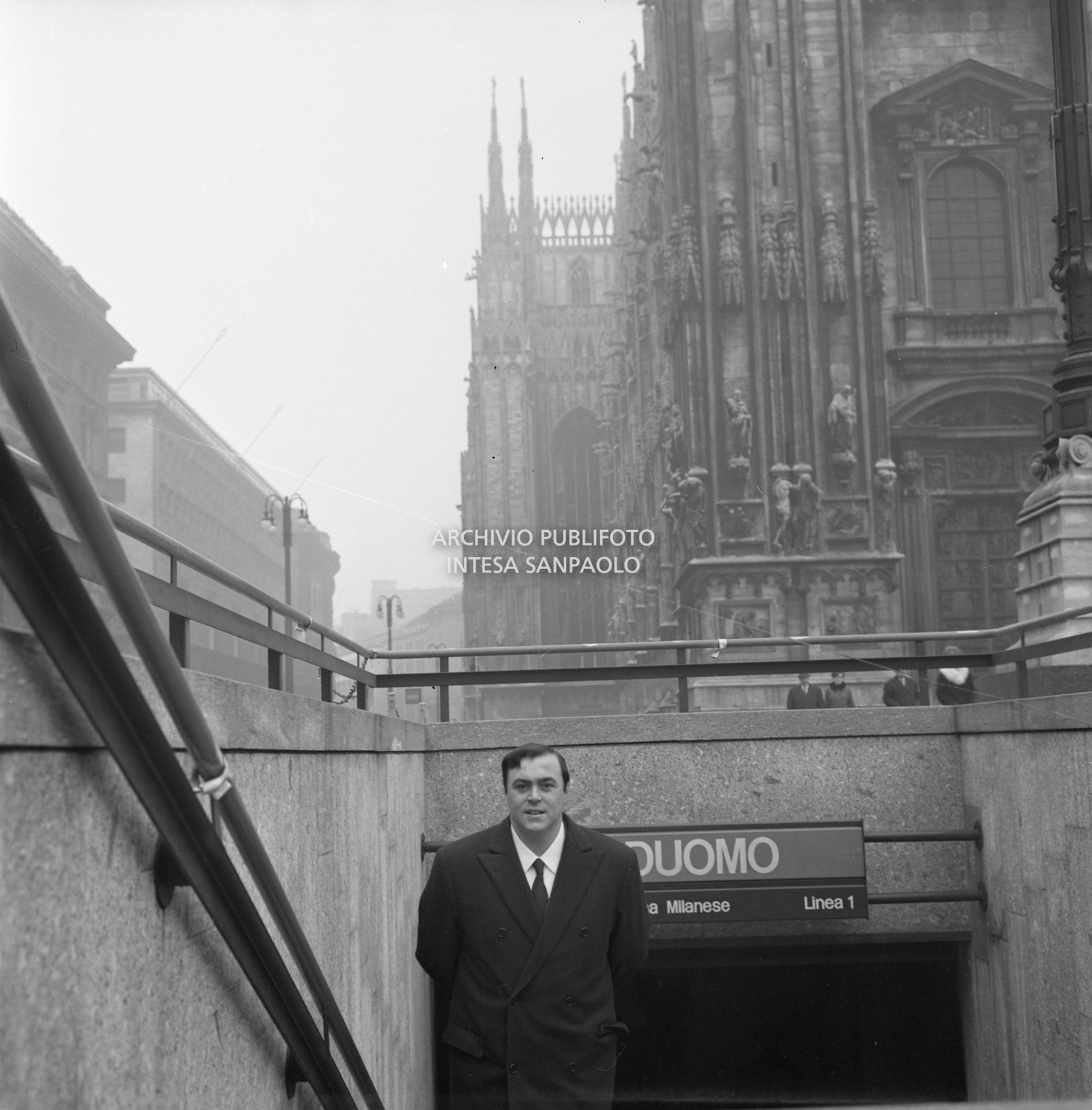 Luciano Pavarotti ritratto sulle scale di ingresso alla stazione Duomo della metropolitana a Milano, sullo sfondo il Duomo